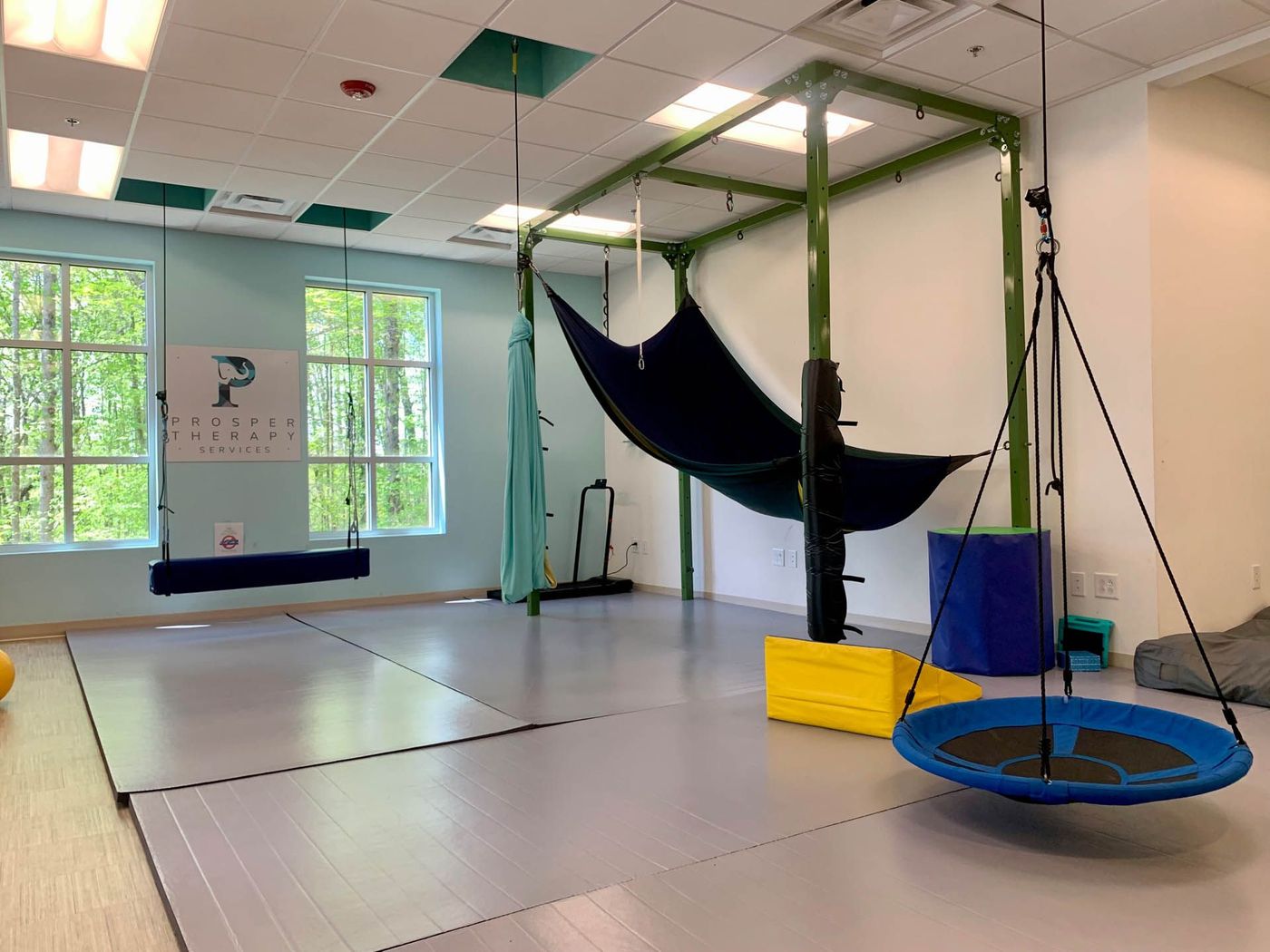 Therapy room with swings, green frame, and blue, yellow, and black equipment. Light blue walls and windows.