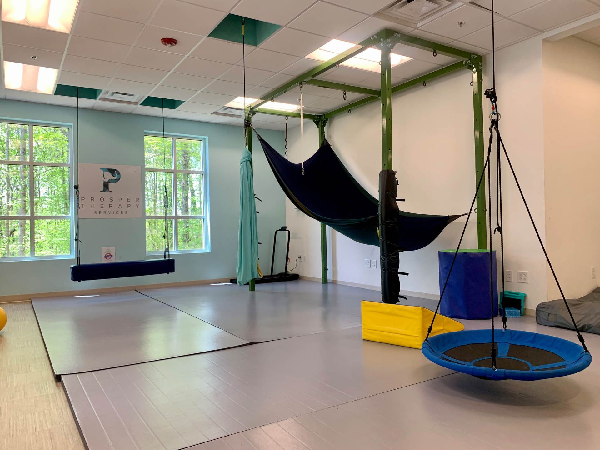Therapy room with swings, green frame, and blue, yellow, and black equipment. Light blue walls and windows.