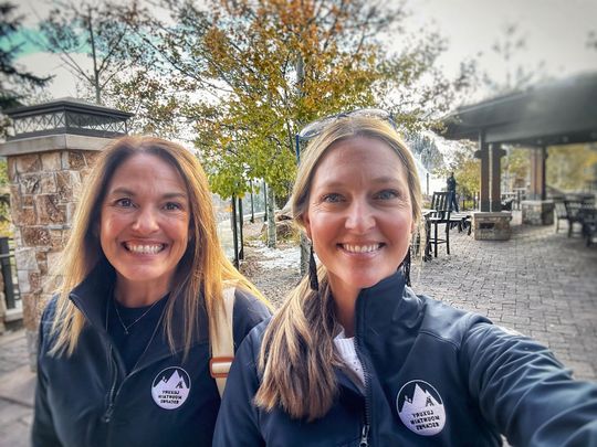 Two smiling people take a selfie outdoors on a sunny patio with trees and stone buildings behind them