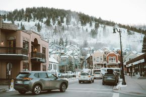 Snowy mountain town street with cars, buildings, and pine-covered hills in winter