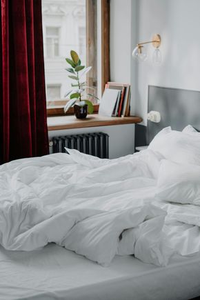 Unmade white bed in a bright bedroom with a window, curtain, plant, books, and wall lamp