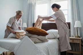 Two caregivers making a bed in a bright bedroom, placing pillows and a blanket on a white bed.