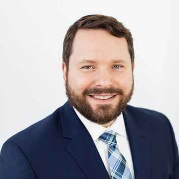 Smiling man with brown hair and beard in a blue suit and patterned tie.