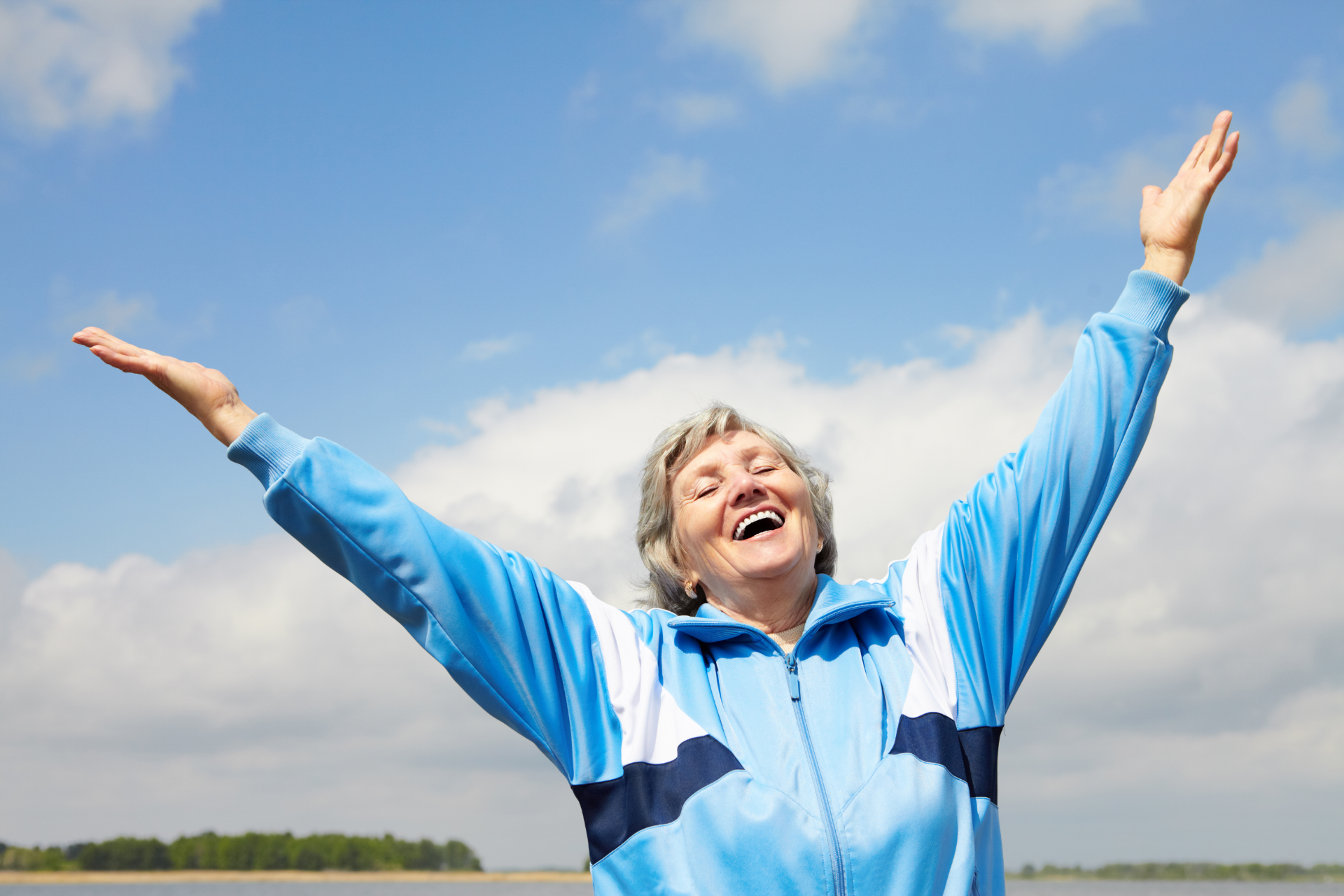 Happy older woman in blue tracksuit raises arms to blue sky.