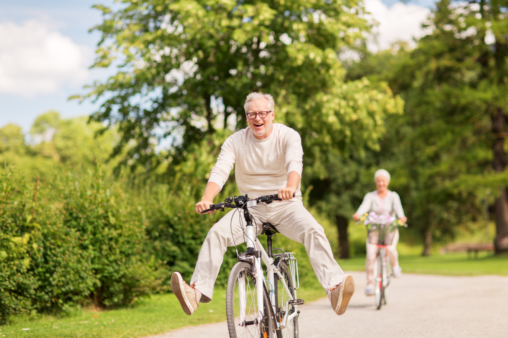 Elderly man joyfully riding a bicycle with legs outstretched in a park, another cyclist behind.