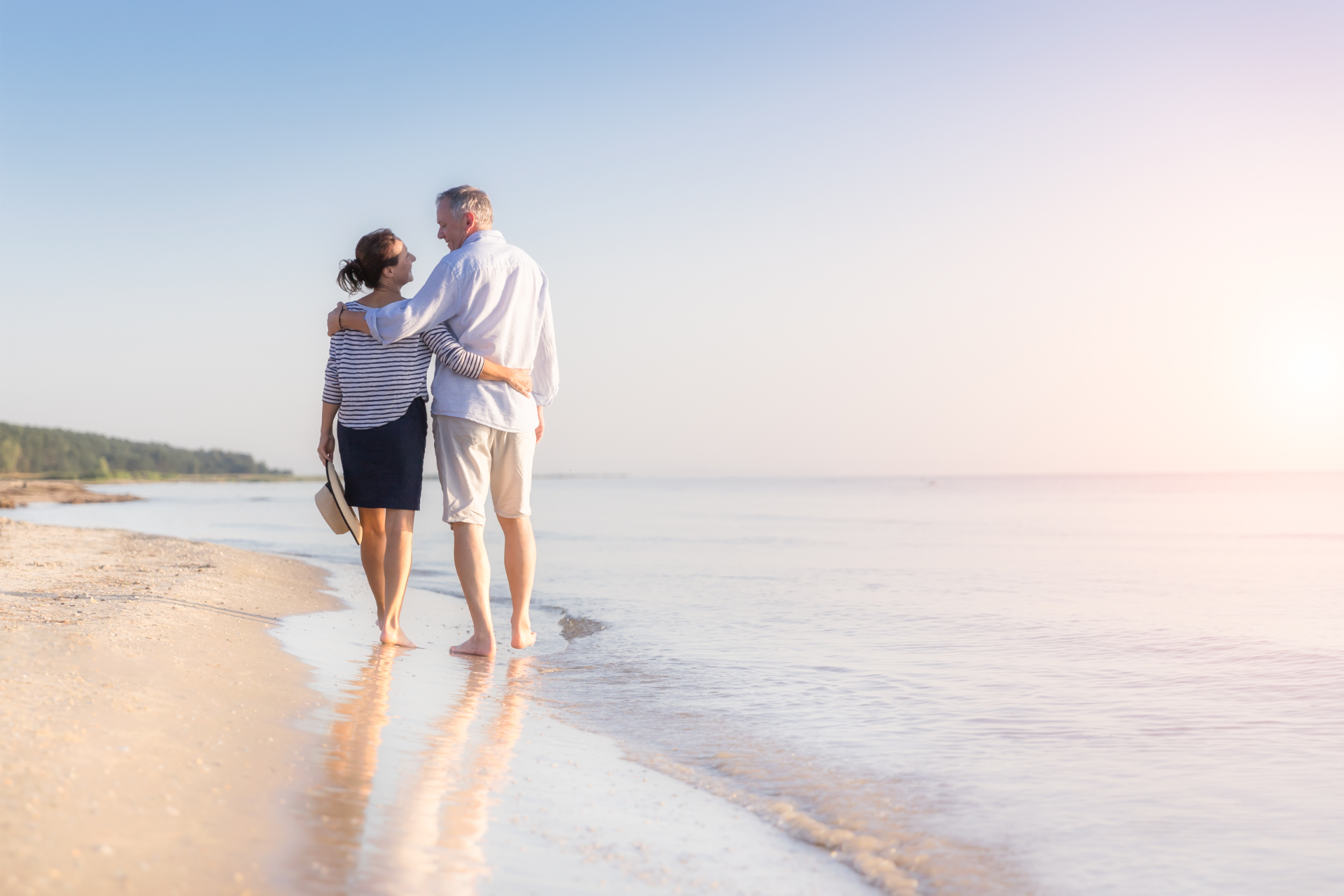 Couple walks along the beach at sunrise, arms around each other, water gently lapping at their feet.
