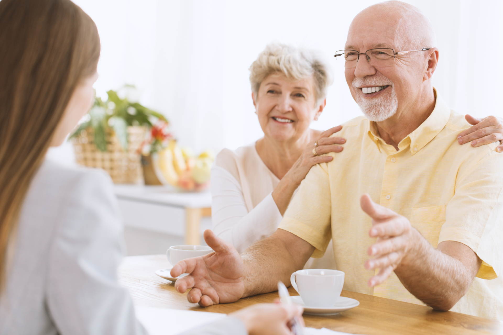An elderly couple smiles, discussing with a professional at a table indoors. The woman has her arm around the man.