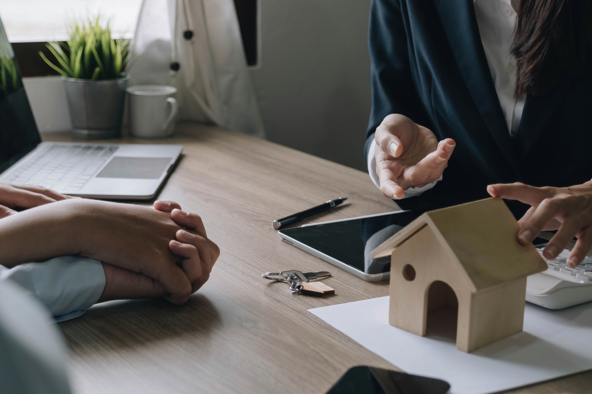 Person points at a model house during a meeting. Laptop, tablet, and keys on the table.