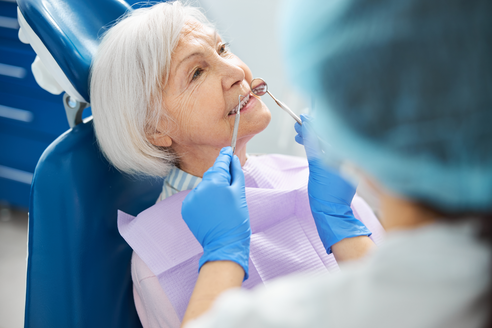 Senior woman in dental chair, receiving dental exam by gloved dentist.