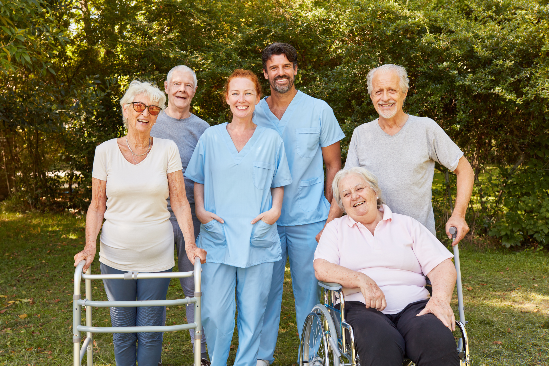 Group of seniors with two caregivers smiling outdoors; some use walkers/wheelchair.