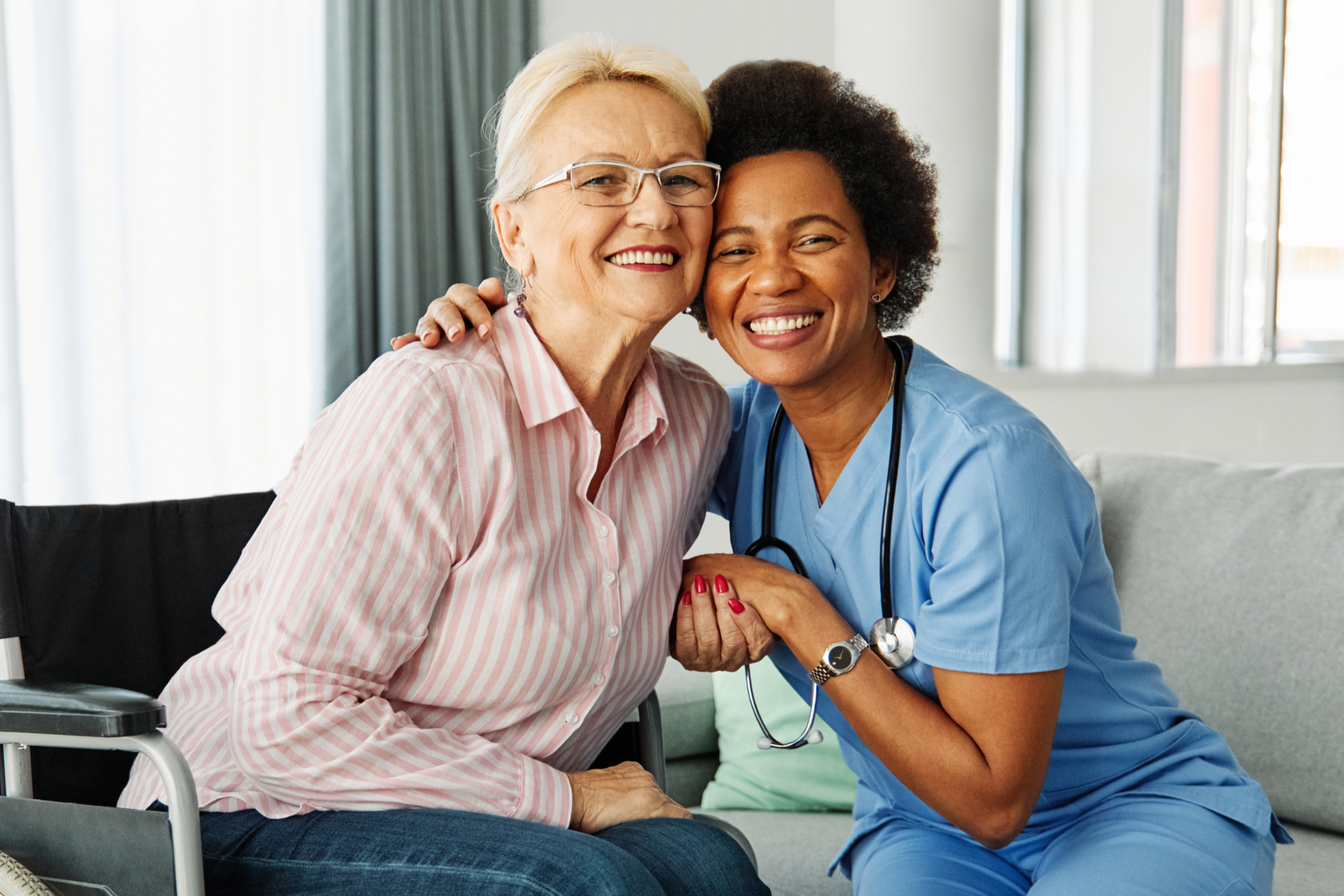 Smiling caregiver hugs an elderly woman, both sitting on a couch indoors.