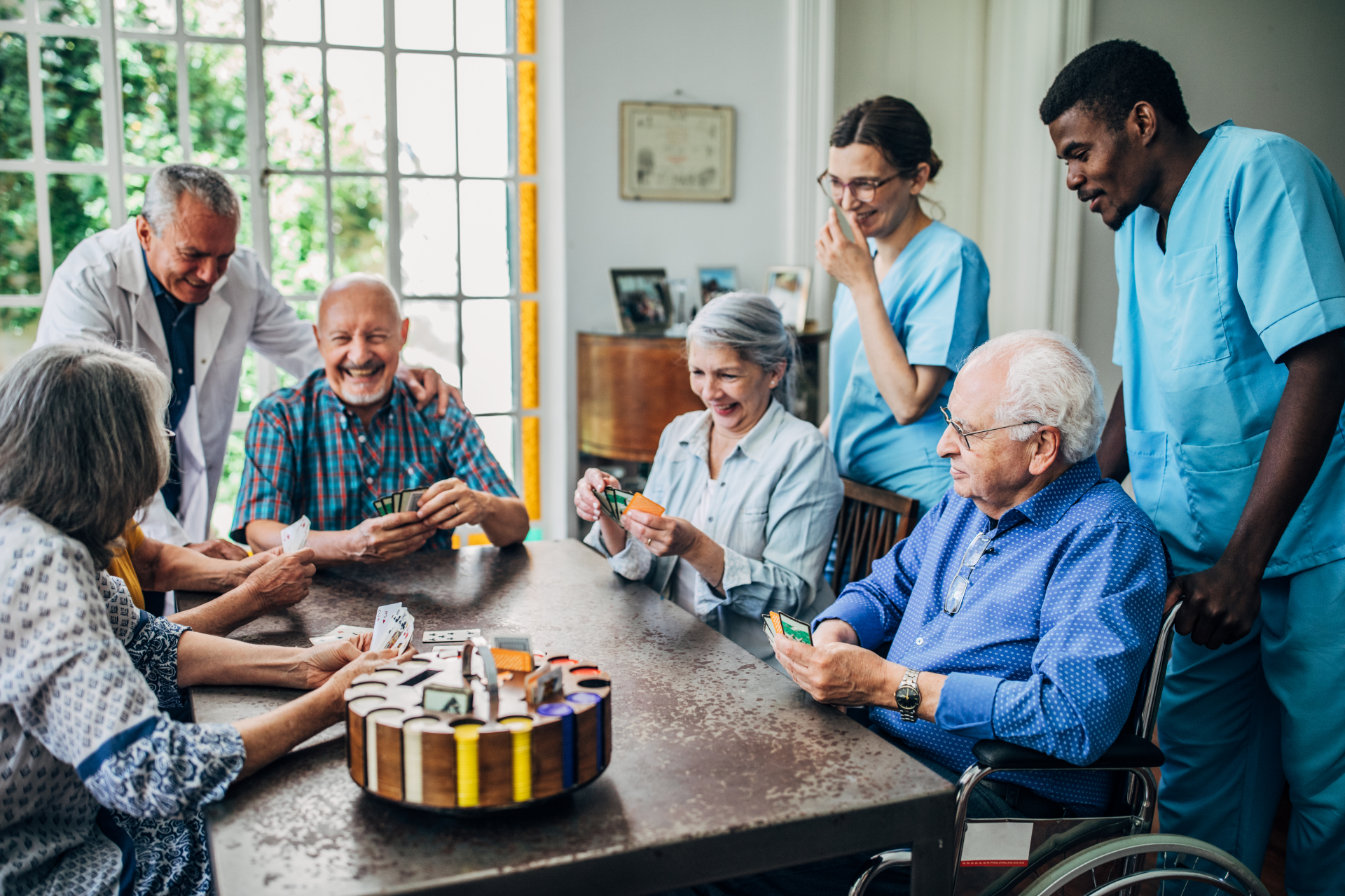 Seniors playing cards at a table, with caregivers observing. Inside a room with large windows.