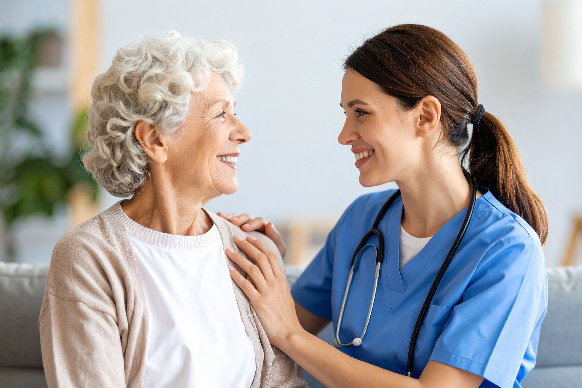 A smiling nurse in blue scrubs comforts an elderly woman. They are indoors on a couch.