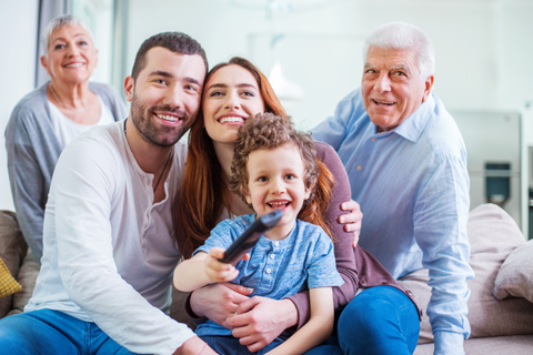 Family of five smiling together on a couch; includes grandparents, parents, and a child holding a remote.