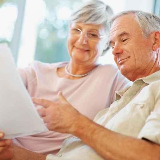 Older couple looking at and pointing to a document. Woman in glasses, man smiling. Indoors, light colors.