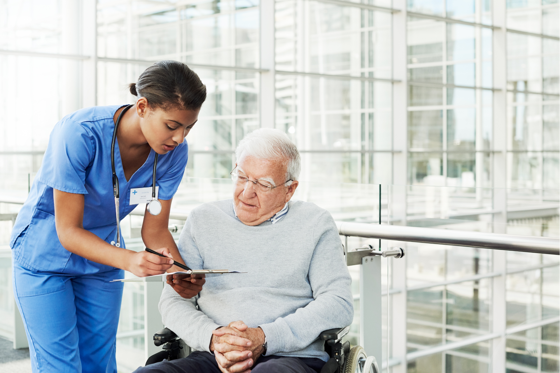 Nurse in blue scrubs reviews paperwork with an elderly man in a wheelchair in a hospital setting.
