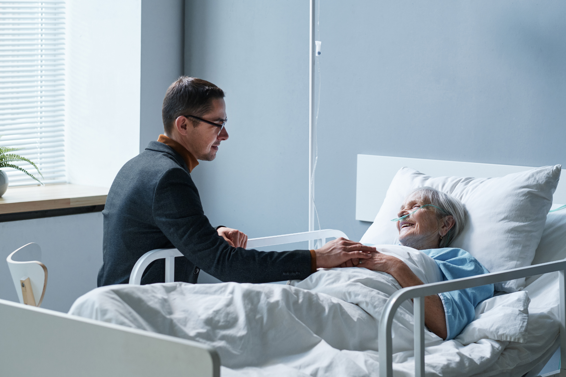 Man comforts elderly woman in hospital bed, holding her hand; window in background.