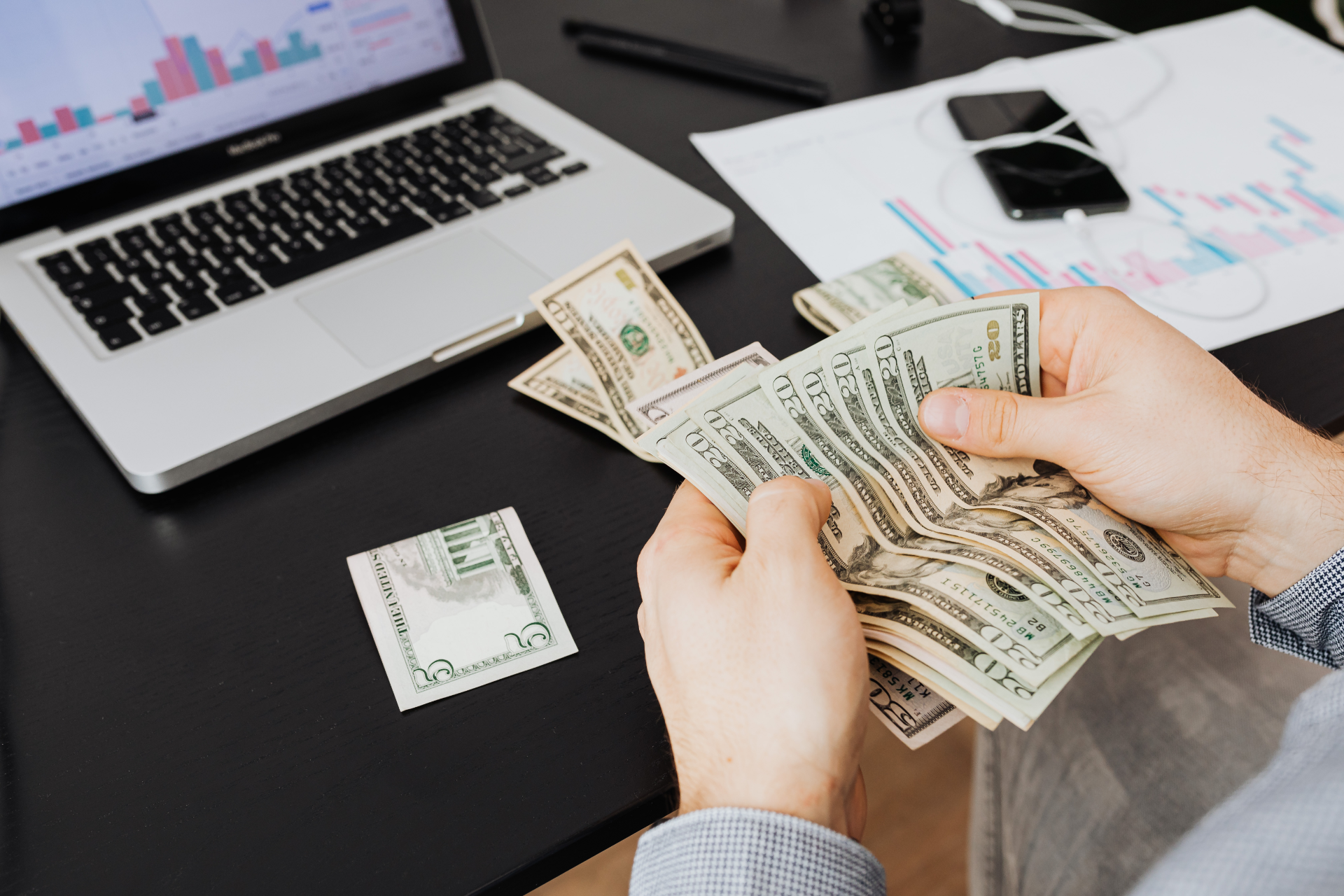 Hands counting cash on a desk with a laptop, charts, and a phone.