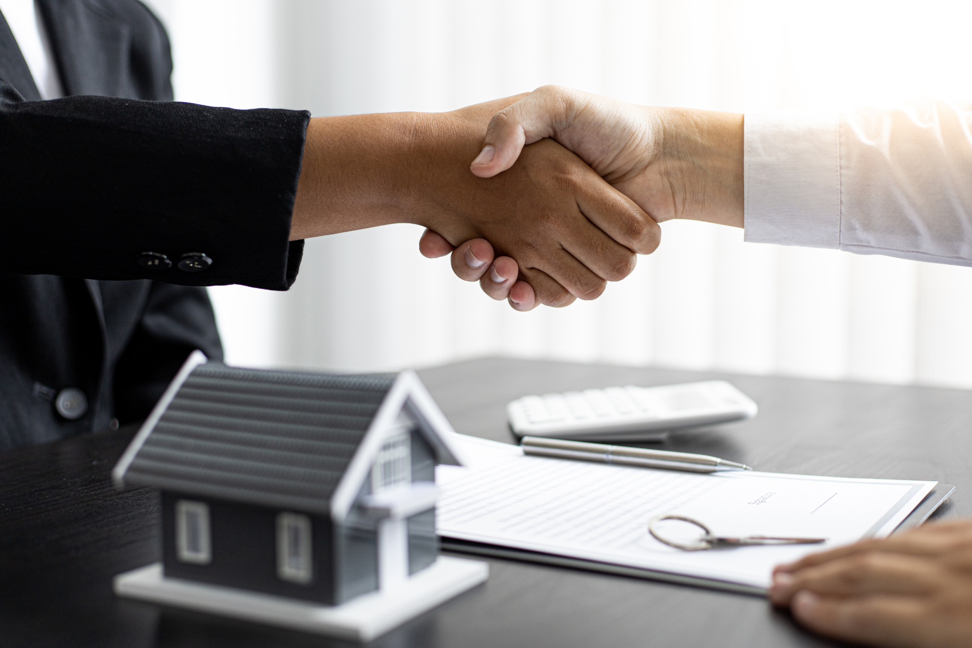 Two people shaking hands over a miniature house, documents, and keys on a table.
