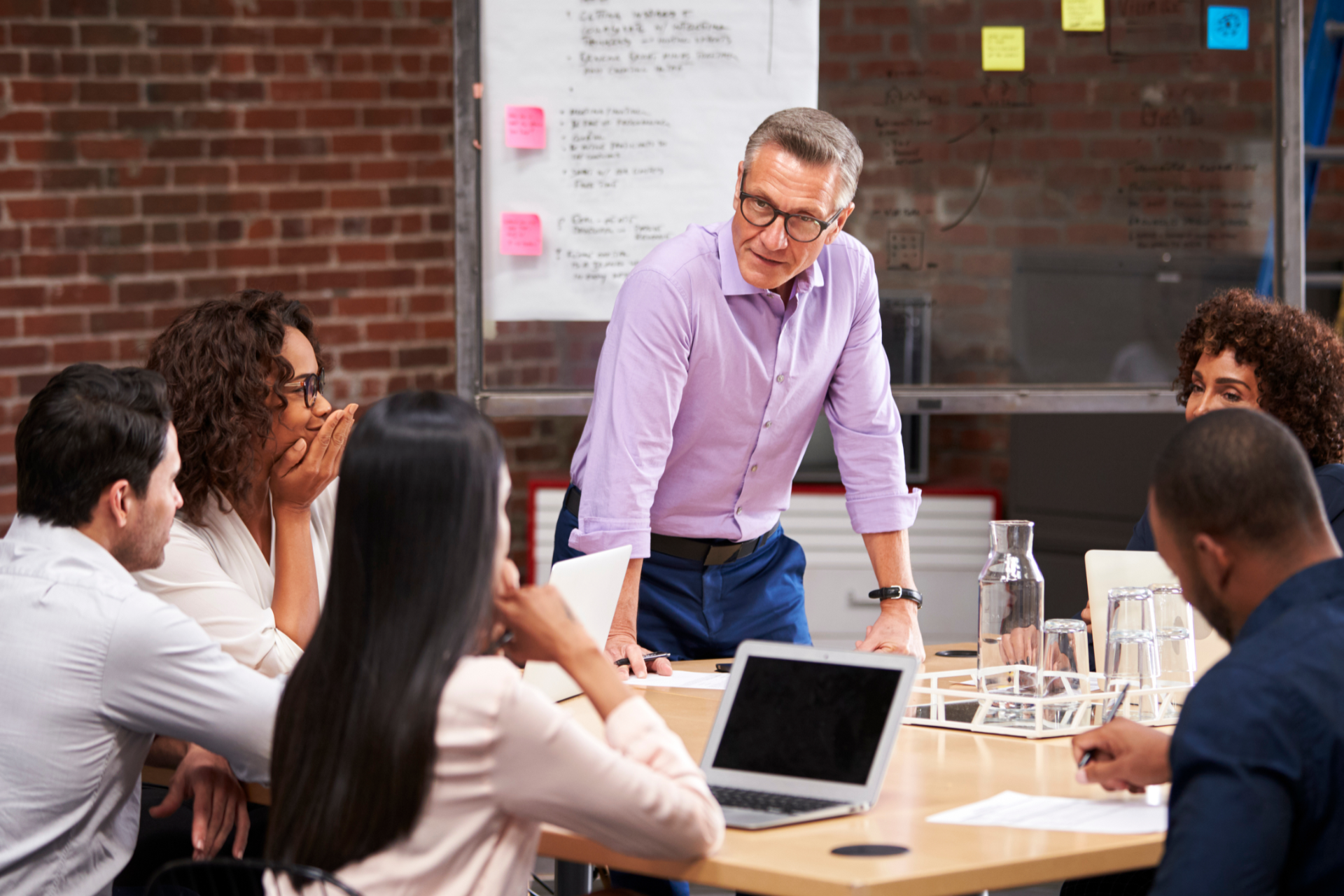 Business meeting: man standing, talking to group around a table in a brick-walled office.