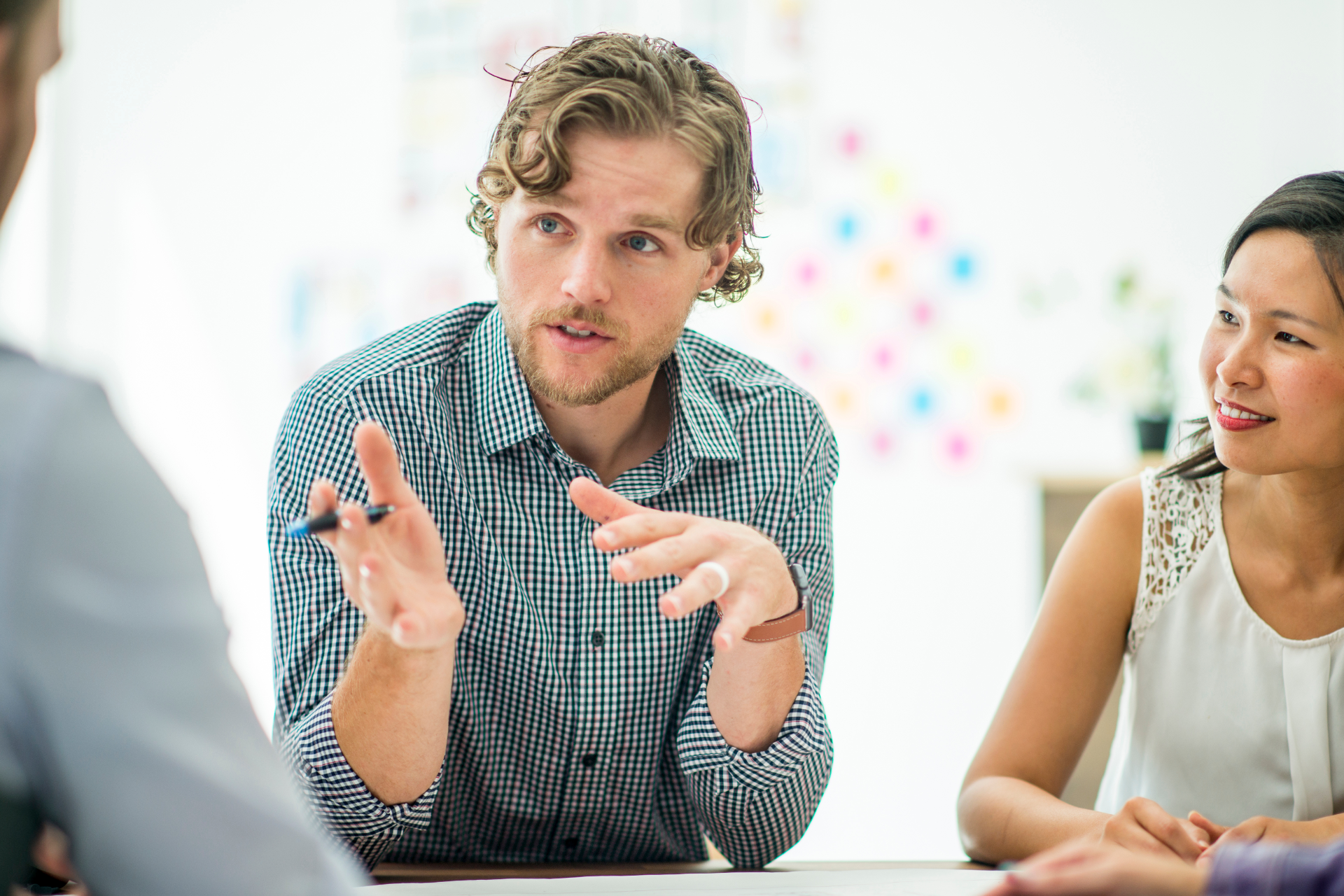 Man with curly hair gestures while speaking to colleagues at a table.