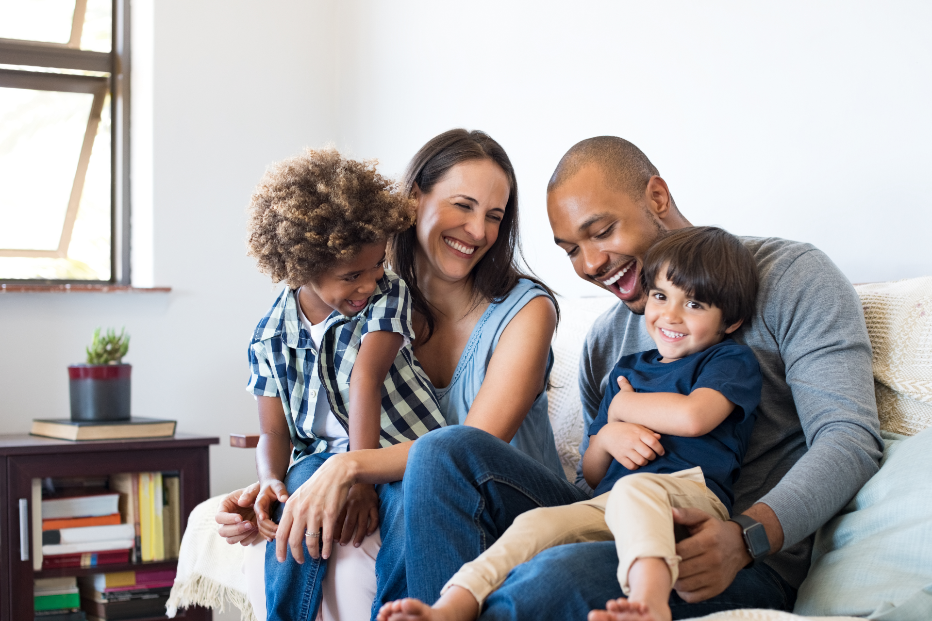 Family of four laughing together on a couch, two children, parents, indoor setting.