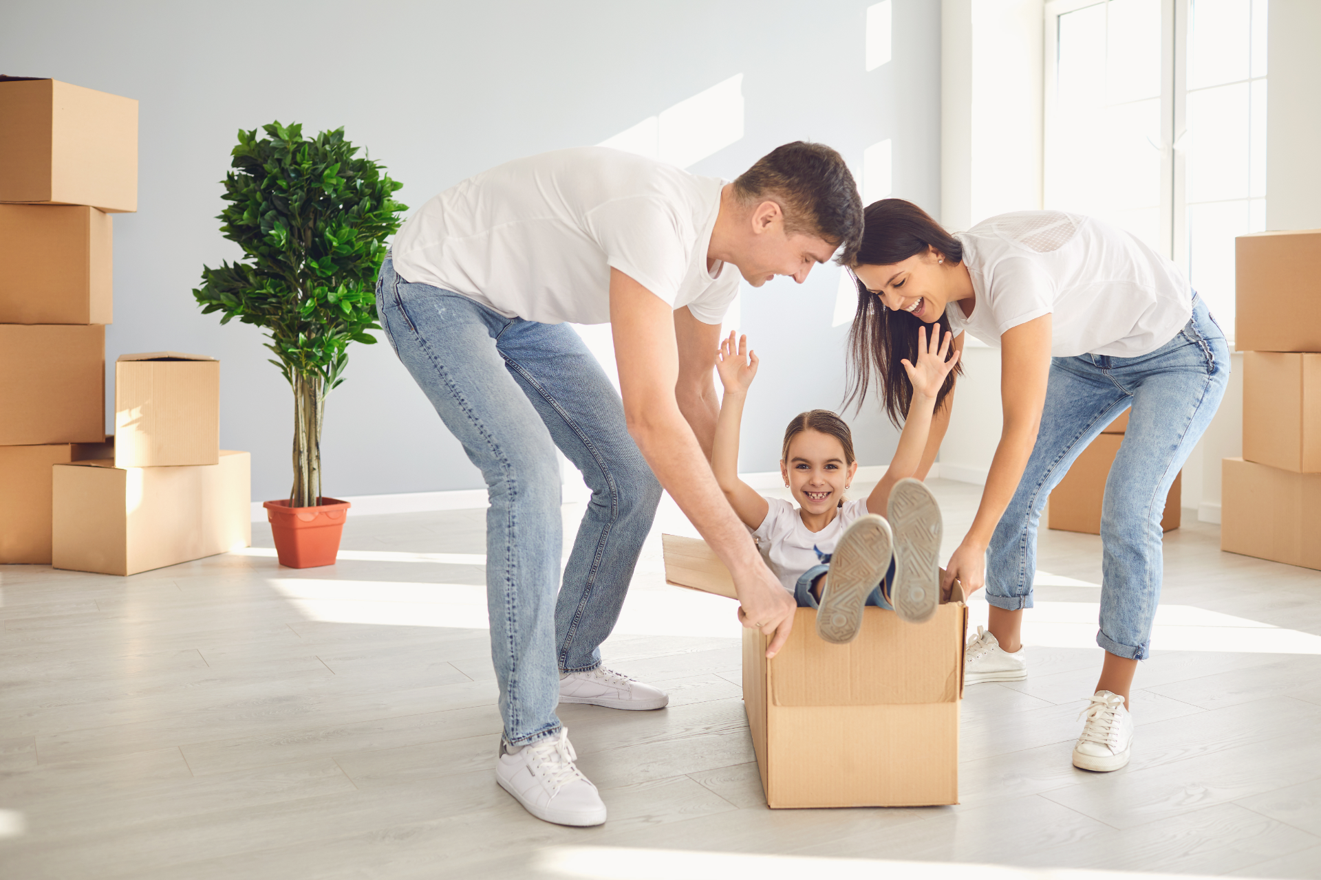 Family playing with daughter in a moving box inside a new, empty home with boxes and a plant.