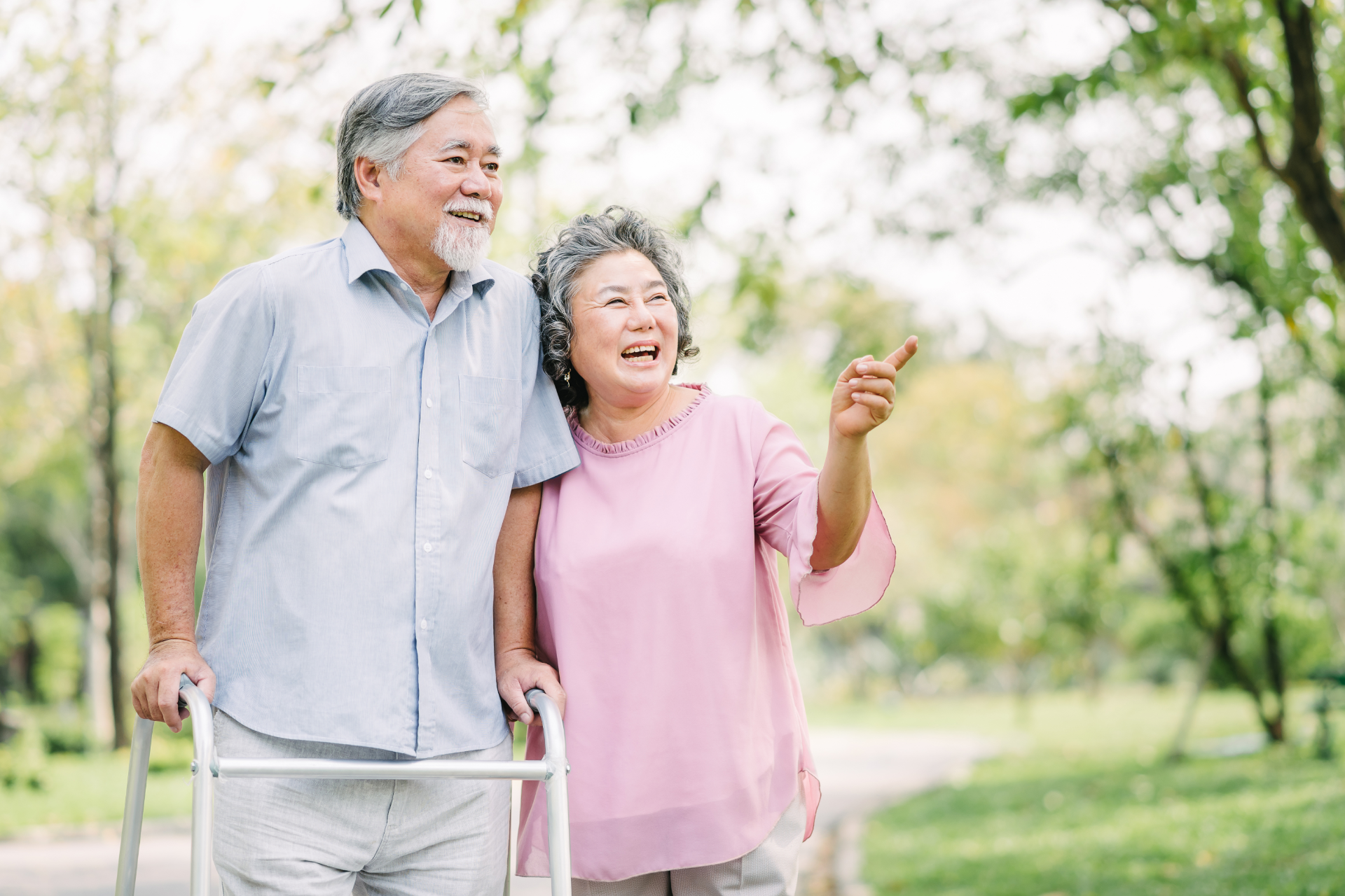 Older couple walking in a park; woman points, man uses a walker, both smiling.