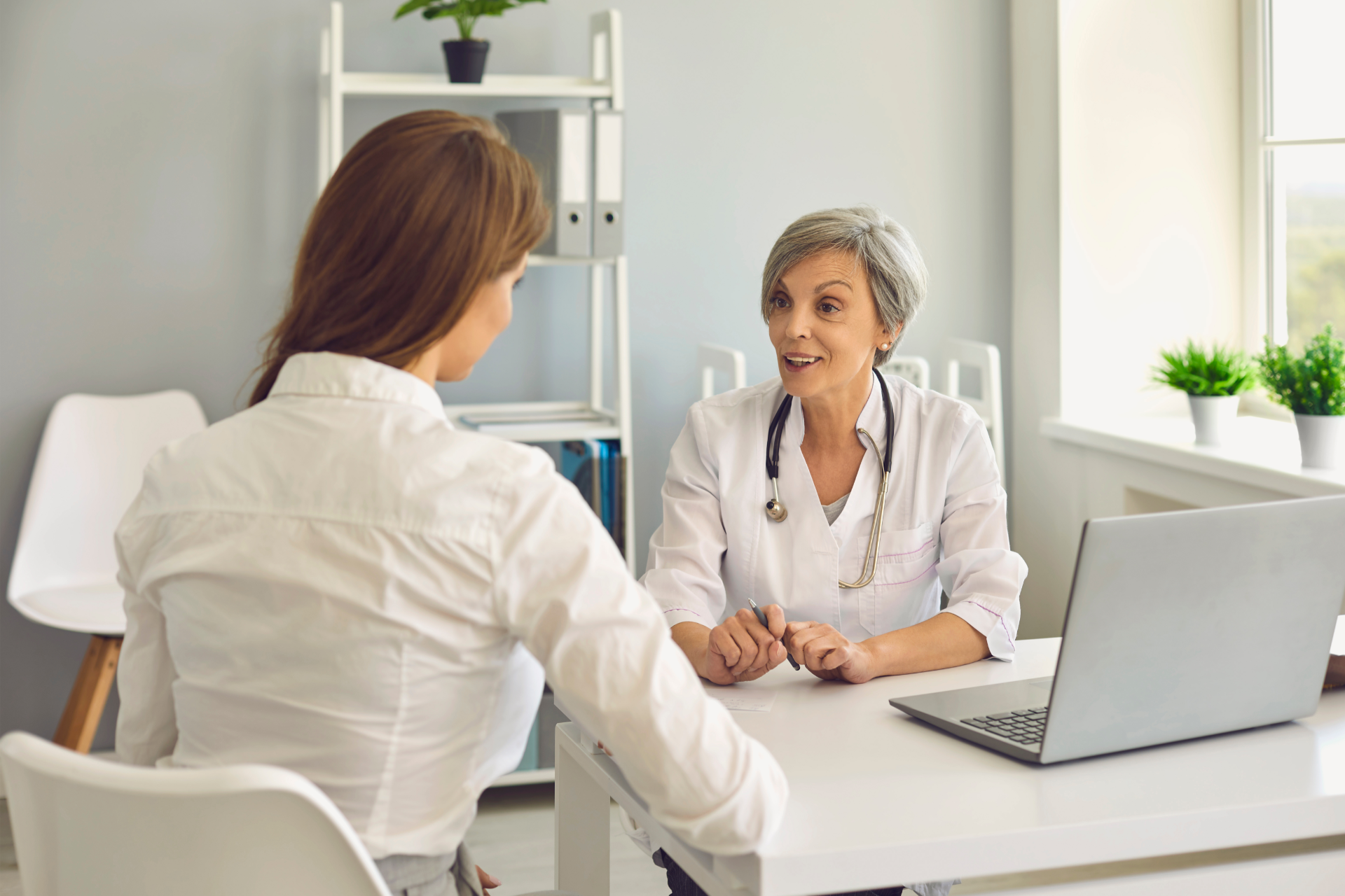 Woman at a desk talking to a doctor with a stethoscope in a medical office.