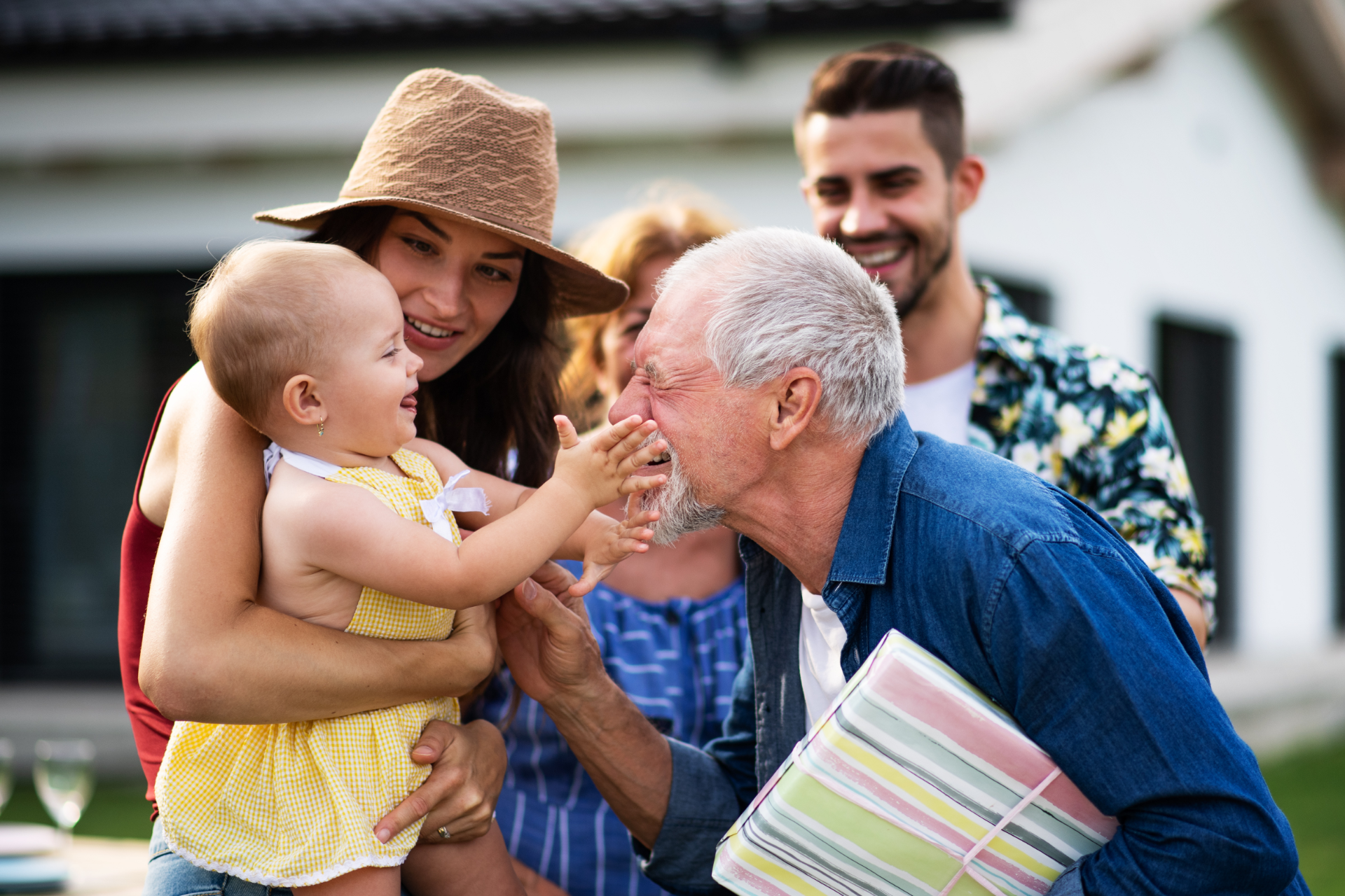 Family gathering outdoors; baby reaching for grandpa's face, surrounded by parents and siblings.