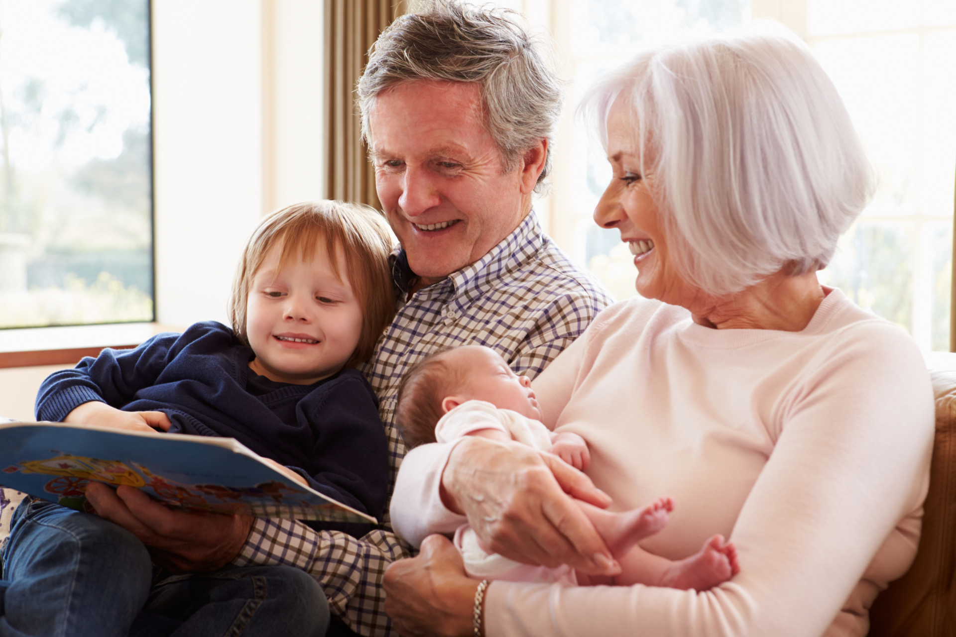 Grandparents reading a book to a toddler and holding a baby, smiling. Indoor setting.