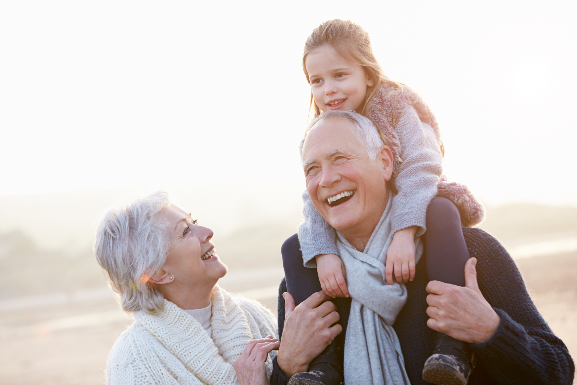 Grandparents with granddaughter. The grandfather is holding the girl on his shoulders, they're smiling. The grandmother is laughing. Outdoors, daytime.