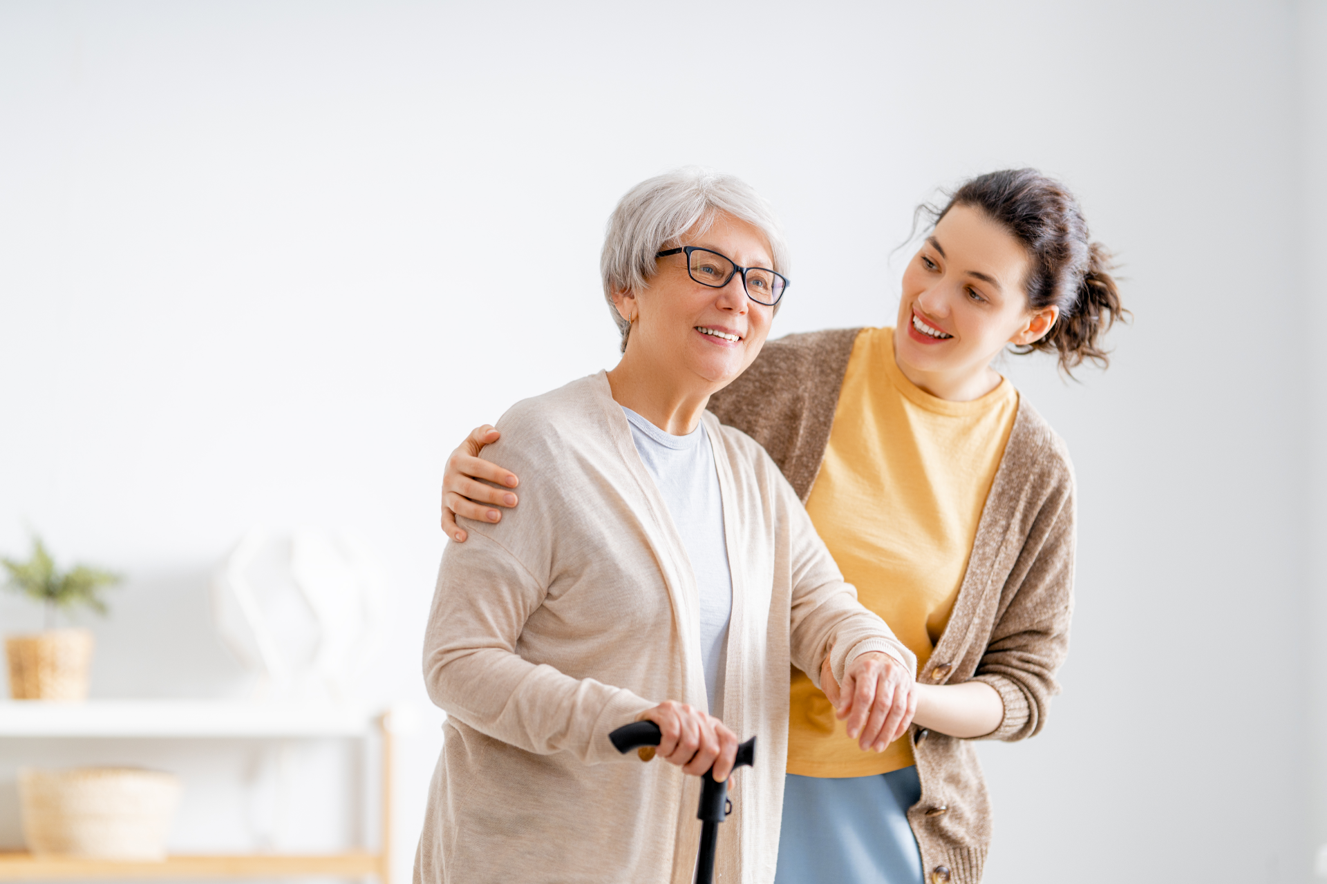 Woman assisting an elderly woman with a cane. Both smile, indoors, white background.