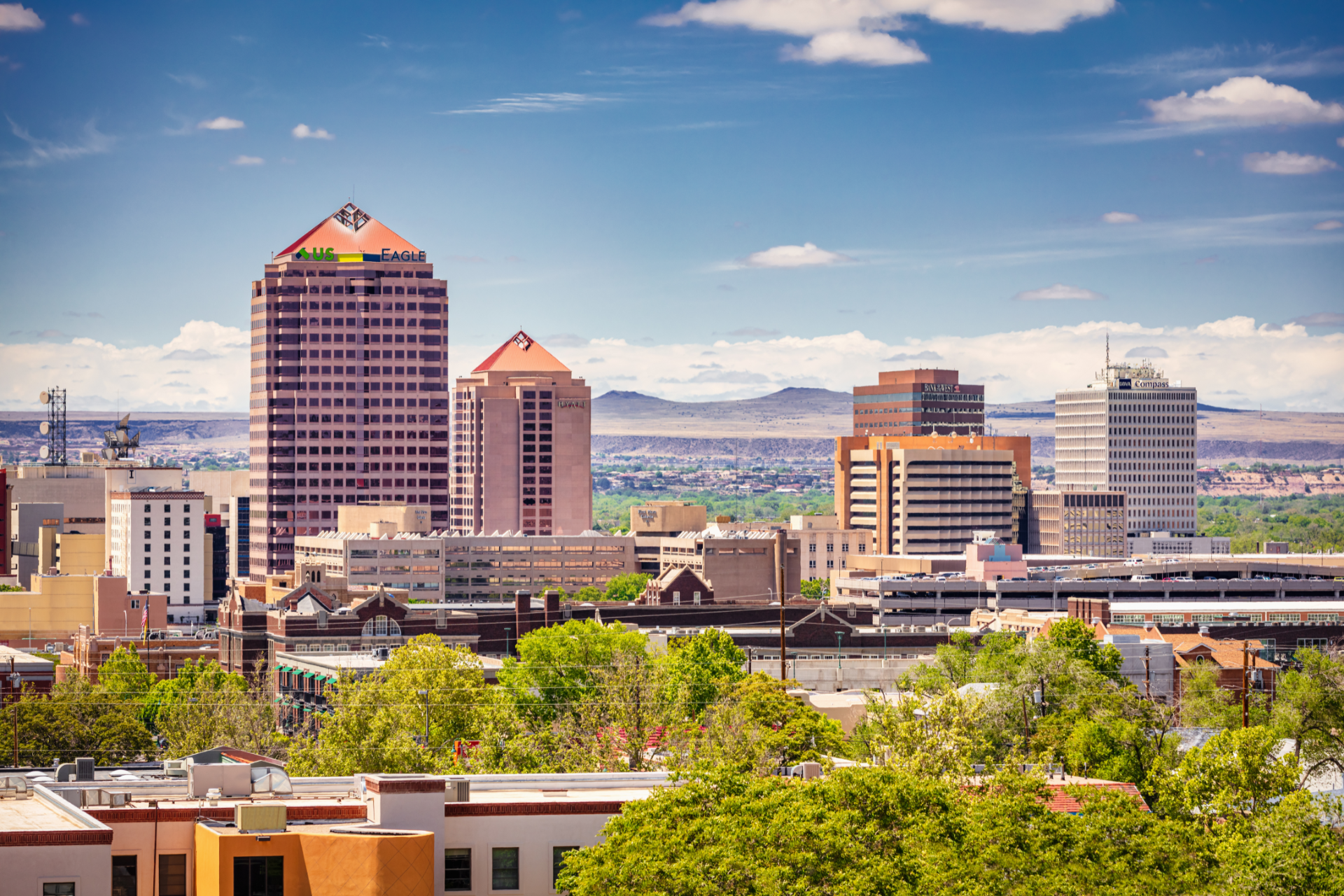 Albuquerque, New Mexico skyline with tall buildings under a blue sky, green trees in foreground, and mountains in the distance.