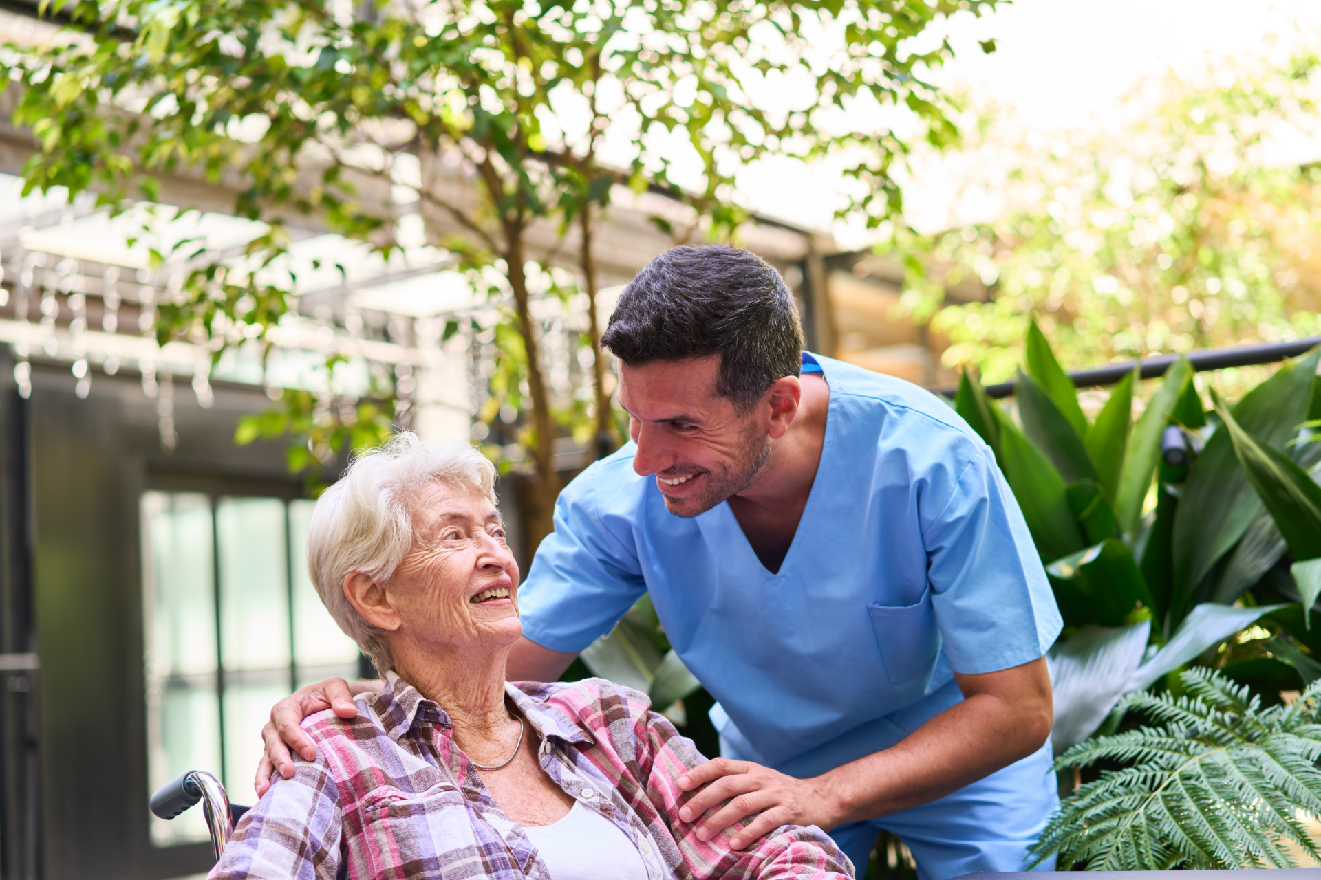 A male caregiver smiles at an elderly woman in a wheelchair, outdoors with greenery.