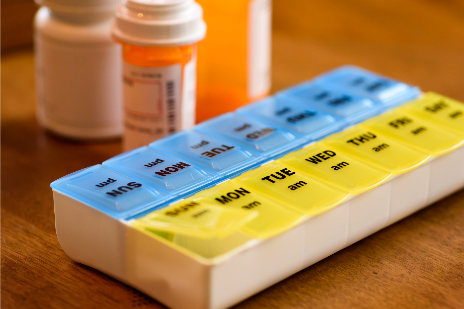Pill organizer with blue and yellow compartments labeled for each day of the week, plus prescription bottles in background.