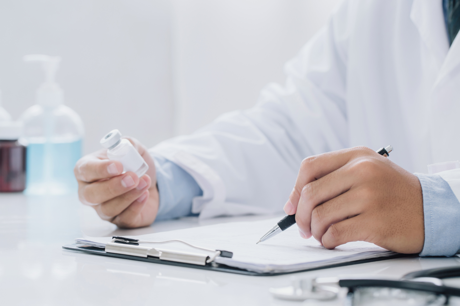 Doctor in white coat holding vial, writing on clipboard with stethoscope, indoors.