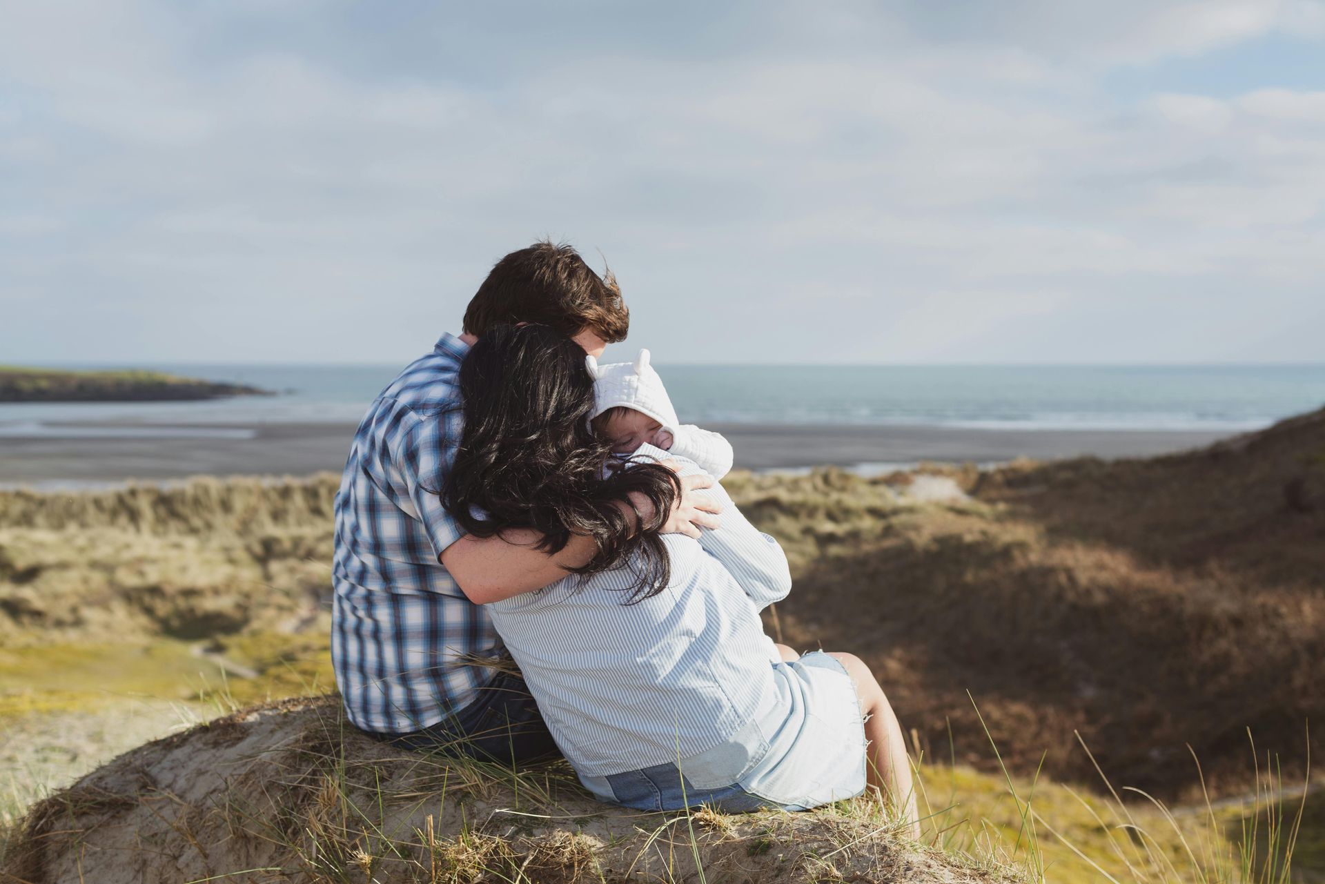 Parents embracing a baby on a cliff overlooking the ocean.