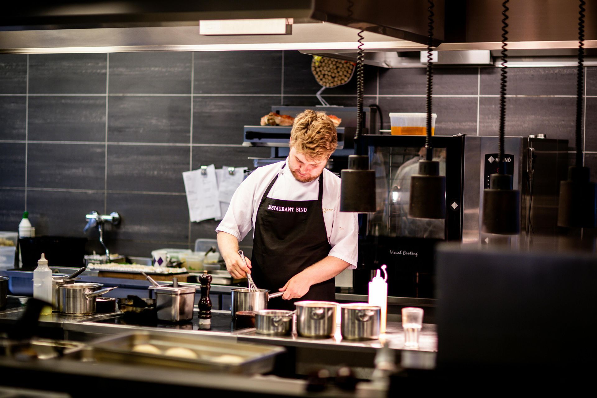 Chef in black apron stirs pots on a stove in a commercial kitchen with stainless steel appliances.