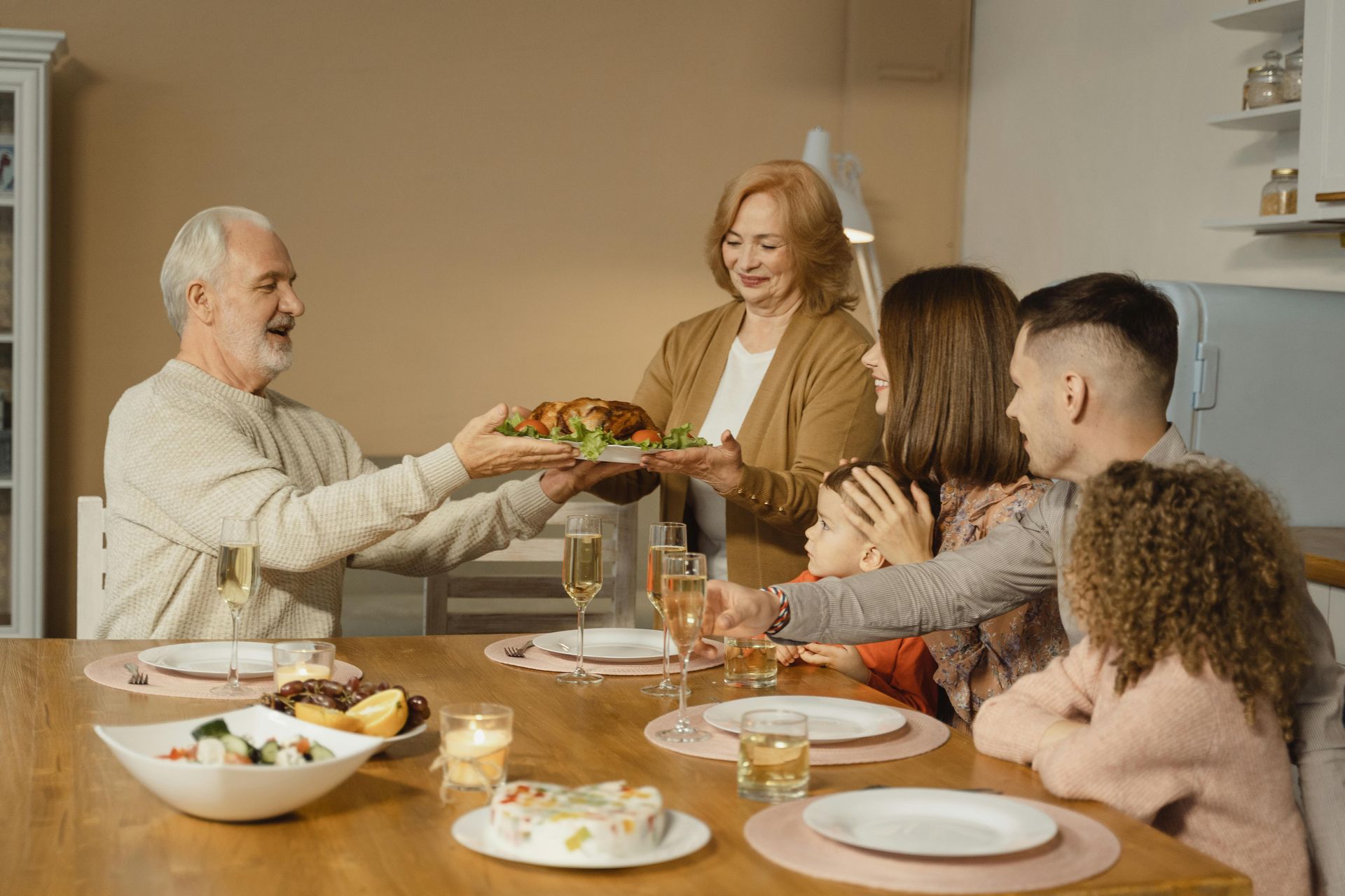 Family gathers around a wooden table; a woman presents a roast chicken.
