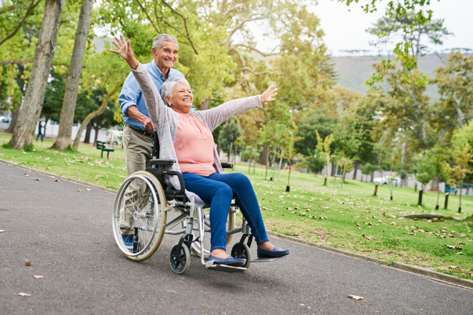 Man pushing woman in wheelchair, both with arms raised joyfully in park.