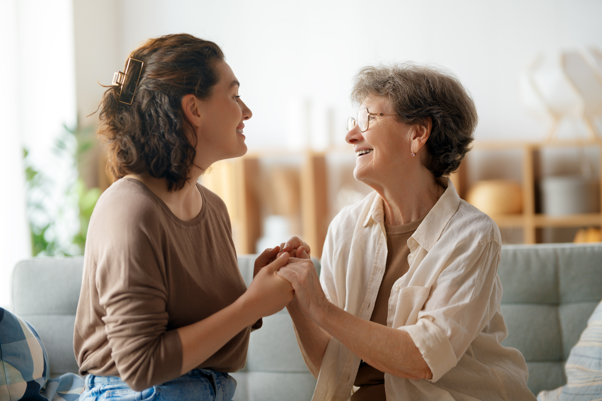 Woman and elderly woman holding hands, smiling, indoors.