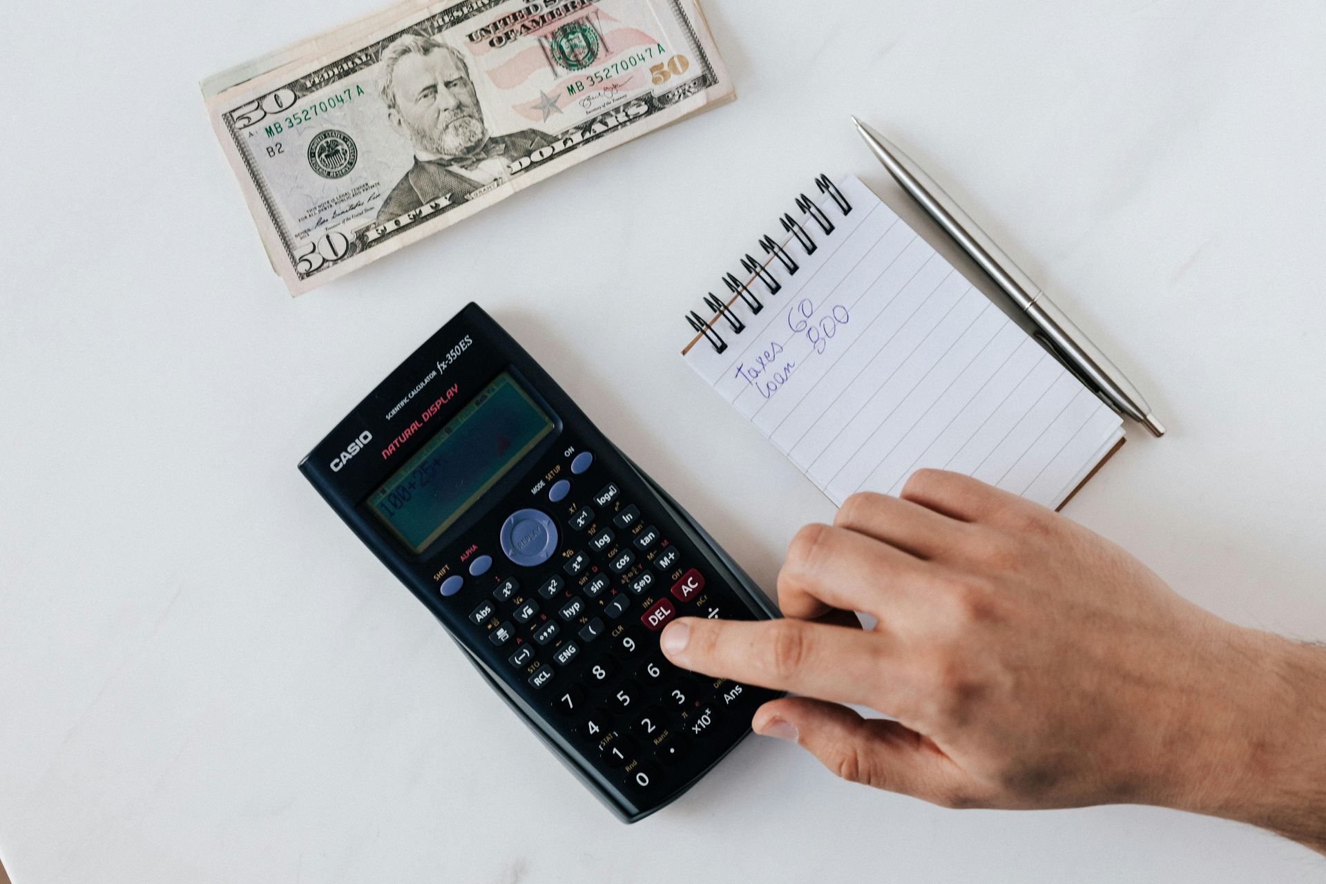 A person is using a calculator next to a notebook and pen.