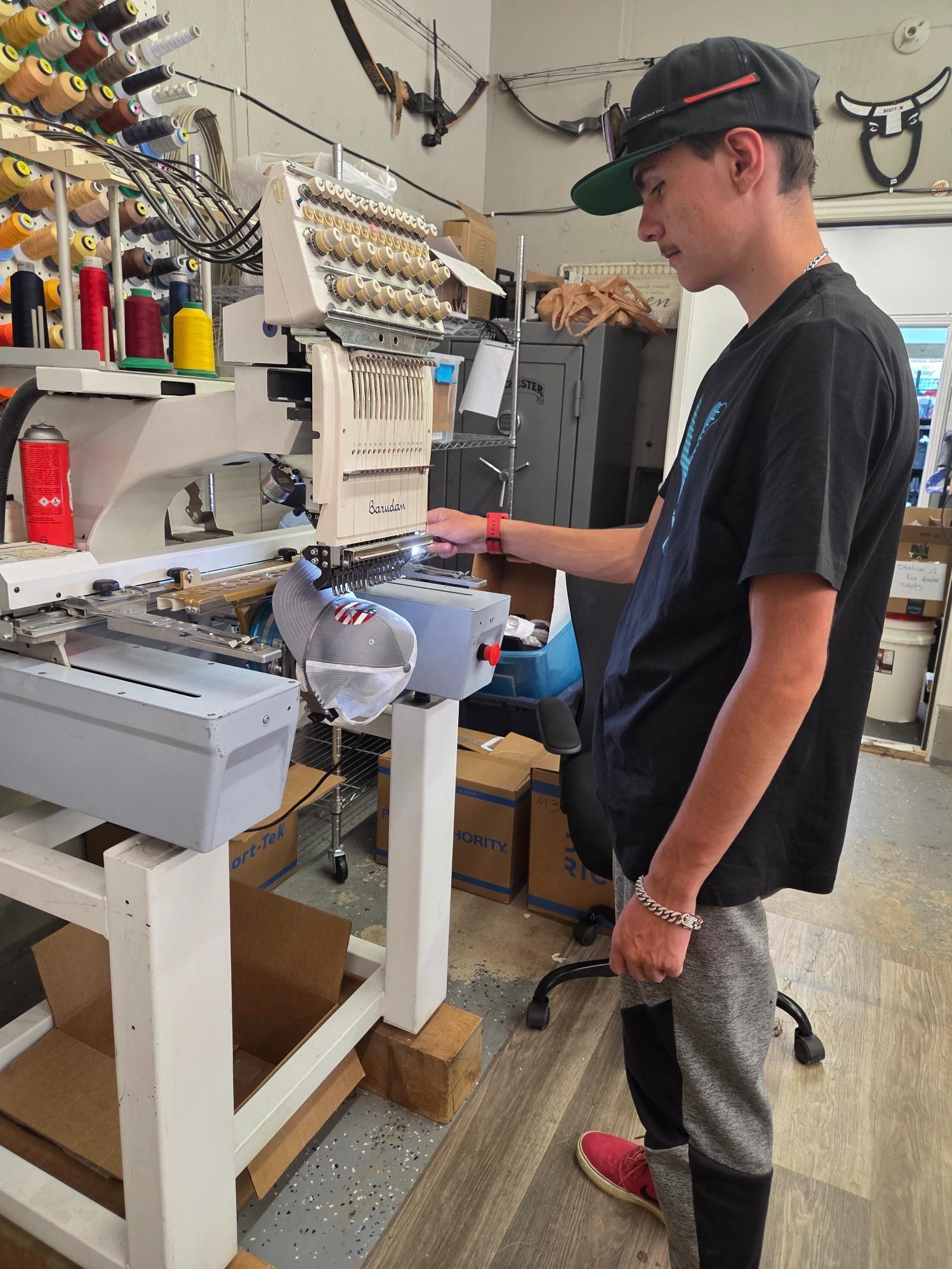 A person operating an embroidery machine in a workshop. The person wears a cap, dark shirt, and is focused.