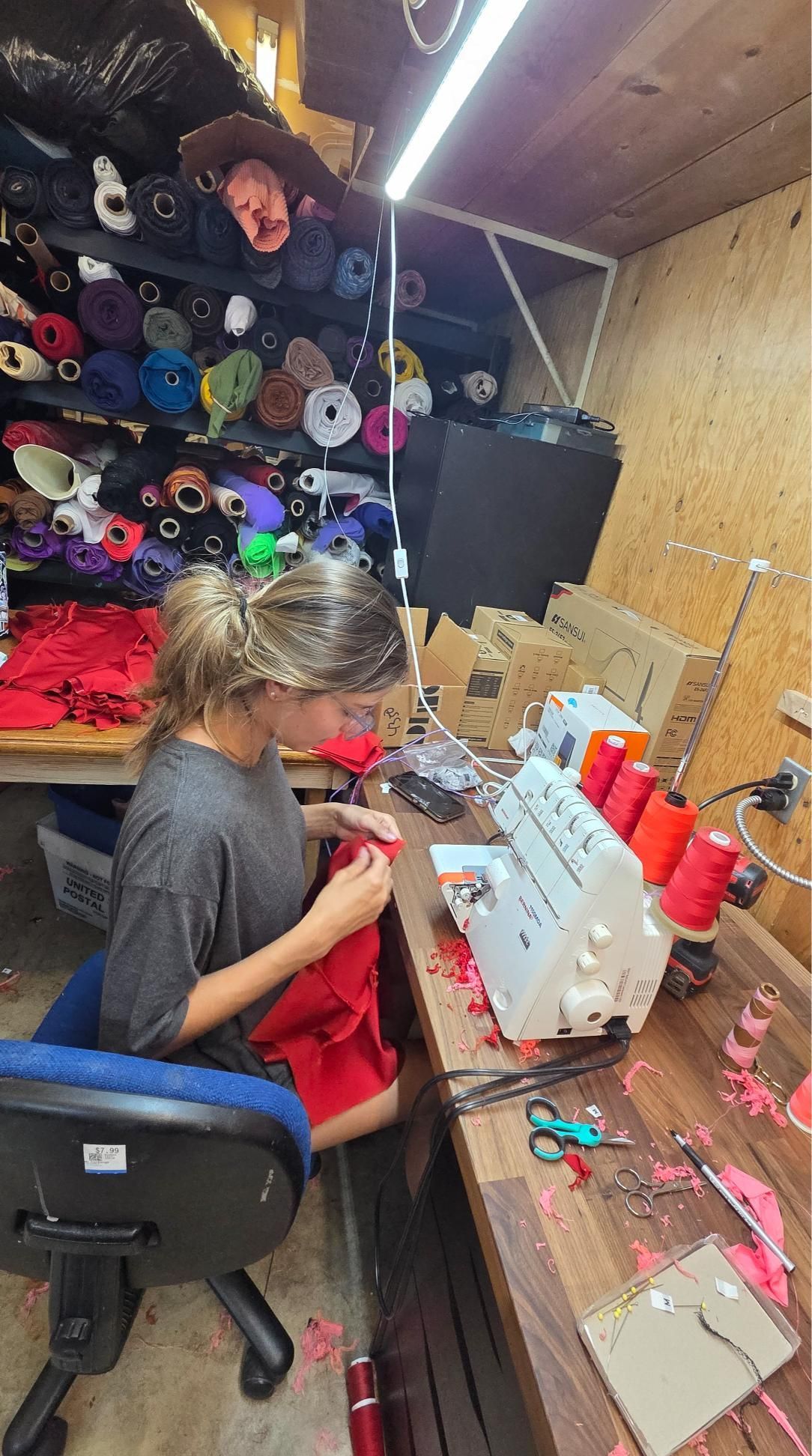 Person sewing at a workbench; rolls of fabric in background. Red fabric and thread. Sewing machine present.