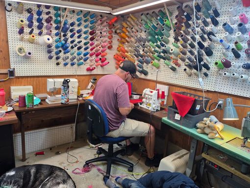 Man sewing at a cluttered workbench, surrounded by spools of colorful thread. Dog rests on the floor.