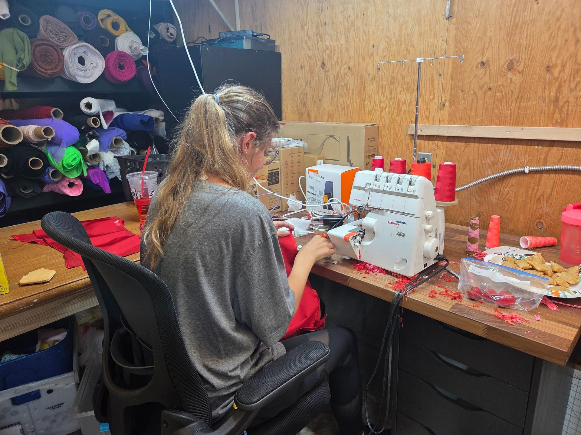 Person sewing at a table with a serger machine, surrounded by fabric and sewing supplies in a workshop.