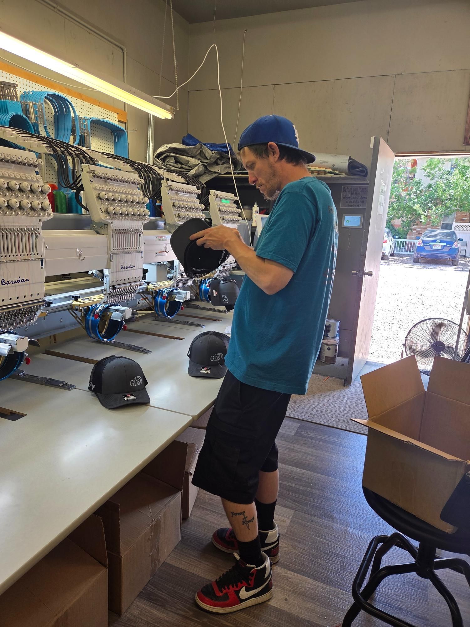 Man in blue shirt and hat examines a black cap in an embroidery workshop, near machines and finished hats.