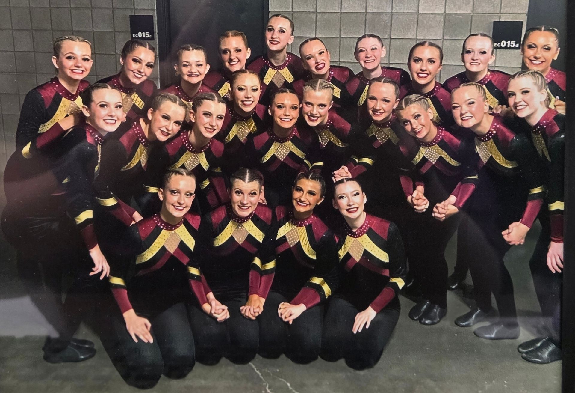 A dance team in black and gold outfits poses together. Dark setting.