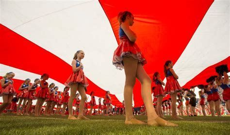 Girls in red and blue dresses hold up a large red and white flag on a green field.